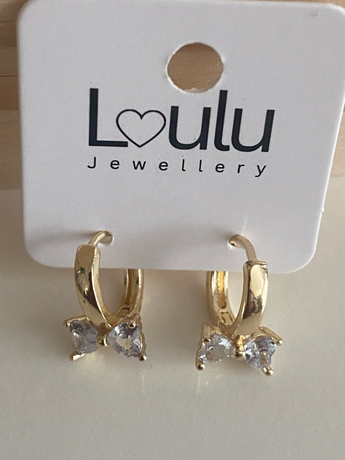 Loulu Jewellery gold-tone huggie hoop earrings featuring a delicate bow motif with sparkling heart-shaped crystals.