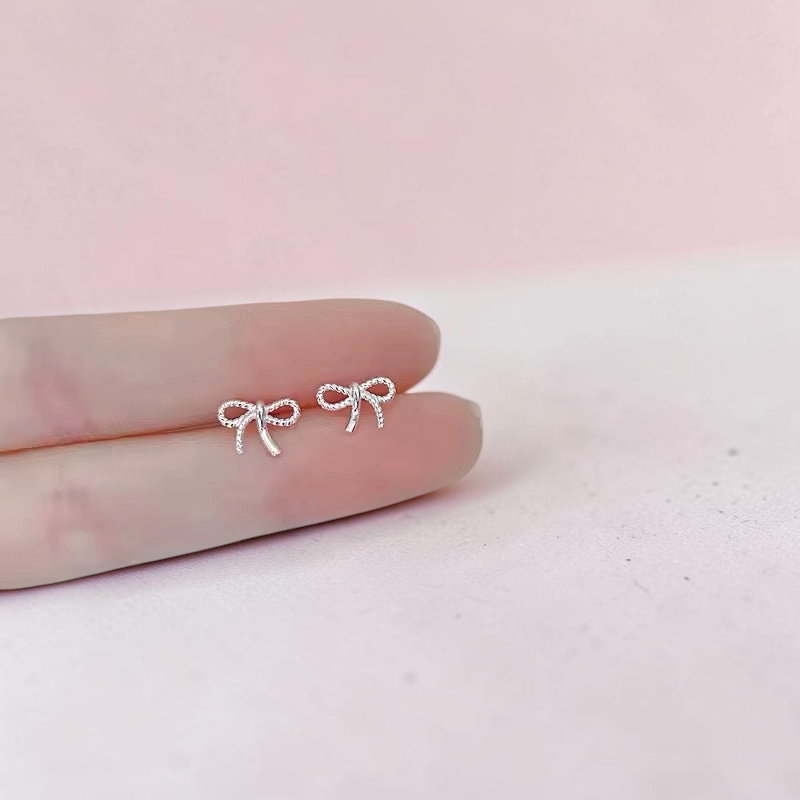 Loulu Jewellery delicate sterling silver bow stud earrings with a twisted rope-effect texture held on a fingertip